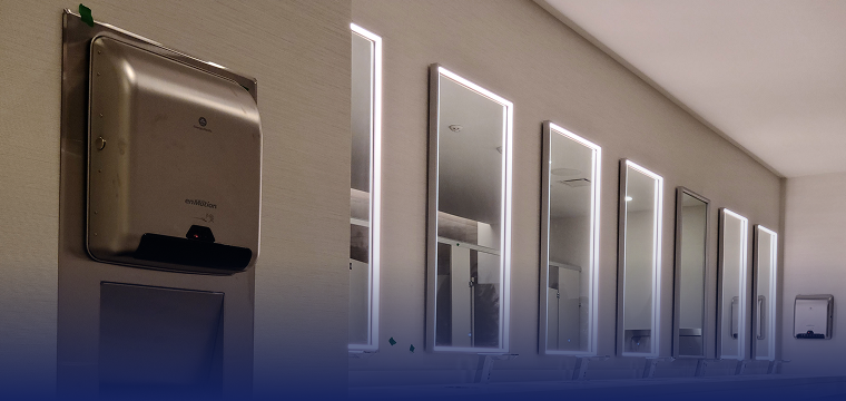 Stainless steel paper towel dispenser beside a row of lighted mirrors in a commercial restroom